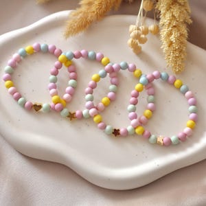 Könnte beinhalten: Vier elastische Armbänder aus matten Pastellperlen in Rosa, Gelb und Hellblau. Jedes Armband ist mit einem einzelnen goldfarbenen Metallanhänger in Form eines Herzens, eines Sterns, einer Blume oder eines Schmetterlings versehen.