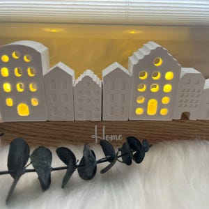 Puede incluir: Una escena de pueblo de cerámica blanca con ventanas iluminadas, montada sobre una base de madera. La palabra "Home" está escrita en la base. Hojas decorativas de eucalipto en primer plano. Los edificios tienen varias formas y tamaños.