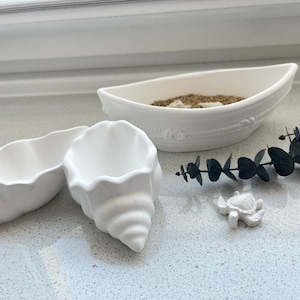 May include: A coastal-themed arrangement featuring two white seashell-shaped containers, a boat-shaped dish with sand, a small turtle figurine, and a sprig of greenery. The items are arranged on a light-colored surface, evoking a beachy aesthetic.