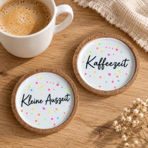 Könnte beinhalten: Zwei runde Untersetzer aus Kork mit weißer Mitte, bunten Konfettipunkten und schwarzem Schriftzug. Ein Untersetzer trägt die Aufschrift Kaffeezeit, der andere Kleine Auszeit. Die Untersetzer liegen neben einer weißen Keramiktasse mit Kaffee.