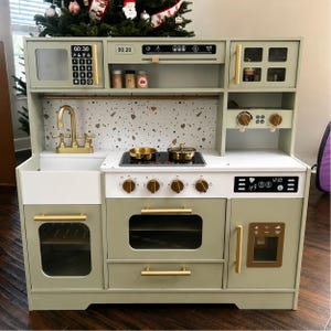 May include: A wooden play kitchen in shades of green and white, featuring a microwave, oven, sink, and refrigerator. It has gold-colored hardware and includes a stovetop with two pots. The kitchen is designed for children.