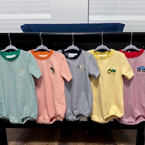 May include: Five striped baby rompers in green, orange, navy, yellow, and red, each with a different embroidered design: a deer, a fish, a bunny, a tractor, and a car. The rompers are hanging on a black surface.