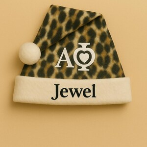May include: A festive Santa hat with a leopard print pattern in shades of brown and black. The hat features a white brim with the word "Jewel" in black letters and the letters "AΦ" in white. A white pom-pom adorns the tip.