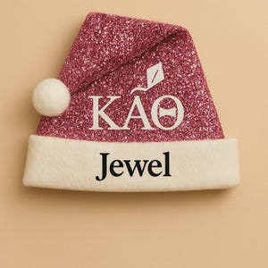 May include: A pink glitter Santa hat with a white pom-pom and trim. The hat features the Greek letters KAO and the word "Jewel" in white lettering. A festive accessory for the holidays.