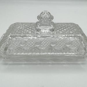 Peut inclure: Un beurrier en verre transparent avec une base rectangulaire et un motif en losange. Le couvercle comporte un bouton décoratif et un motif de volutes répétées. Le plat est conçu pour contenir du beurre.