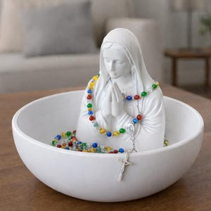 Puede incluir: Una estatua de cerámica blanca de la Virgen María con un rosario colorido y un crucifijo plateado, colocada dentro de un cuenco blanco. La estatua está en una pose de oración. El cuenco está sobre una mesa de madera.