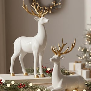 Può includere: Due statuette di renne in ceramica bianca con corna e zoccoli dorati. Una statuetta è in piedi, l'altra è sdraiata. Verde festivo e luci circondano le renne, suggerendo un tema natalizio.