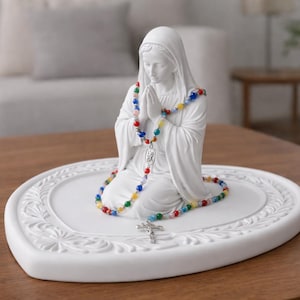 Pode incluir: Estátua de cerâmica branca de uma figura ajoelhada com as mãos juntas em oração. A estátua é adornada com um rosário de contas coloridas e repousa sobre uma base em forma de coração. Uma cruz prateada pende do rosário.