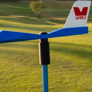 May include: A blue and white Whirly wind spinner with a red V logo on a white background. The wind spinner is on a blue pole with a black connector. The background is a green golf course with trees and a blue sky.