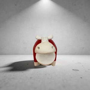 May include: A red and cream-colored cow figurine with a minimalist design. The cow has a red body, a cream-colored face, and small, white ears. The figurine is set against a gray background and is illuminated by a single overhead light.
