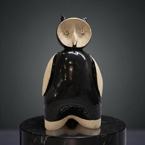 Może przedstawiać: Ceramiczna figurka sowy z czarnym korpusem i kremową głową i akcentami. Sowa ma małe, spiczaste uszy i stylizowaną twarz. Figurka jest eksponowana na czarnym marmurowym cokole.