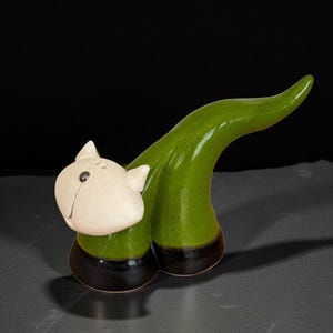 May include: A ceramic cat sculpture with a white head and green body. The cat has a curved tail and a dark brown base. The sculpture has a glossy finish. The design is whimsical and stylized.