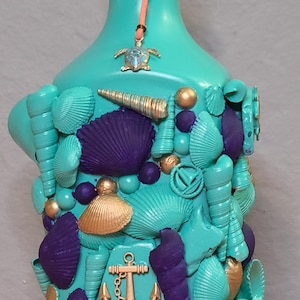 May include: A turquoise bottle decorated with seashells, gold accents, and a purple seashell. The bottle has a cork stopper and a turtle charm. The bottle is decorated with an anchor and other nautical elements.