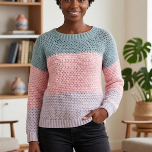 May include: A colour block crochet sweater with blue, pink, and lavender stripes. The long-sleeved sweater has a round neckline. The person in the image is wearing the handmade garment.