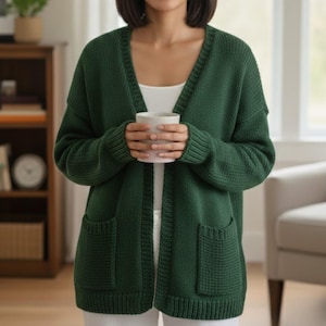May include: A dark green, open-front cardigan with two front pockets. The sweater has a V-neck and a textured knit pattern. The person in the image is holding a white mug. The cardigan is a comfortable, casual style.