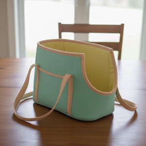 May include: A mint green pet carrier with a light peach trim and straps. The interior is yellow. The carrier has a rectangular shape with a curved opening and a small pocket on the side. The straps are long enough to be worn over the shoulder.