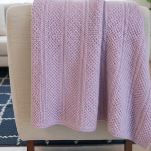 以下が含まれることがあります： ベージュのアームチェアにかけられたラベンダー色のニットのスローケット。ブランケットは、垂直のリブ編み部分とドット模様のテクスチャードパターンが特徴です。柔らかく居心地の良いブランケットは、部屋に彩りと暖かさを加えます。