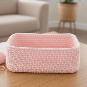 May include: A light pink, rectangular crochet basket sits on a wooden table next to a ball of yarn. The basket is made of a tightly woven stitch and has rounded corners. The yarn is the same color as the basket. The background includes a beige armchair and a plant.