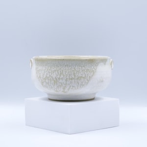 May include: A ceramic bowl with a textured, off-white glaze. The bowl has a rounded shape with two small handles. It sits on a white square pedestal against a light background. The bowl's rim has a slightly darker hue.