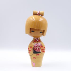 Pode incluir: Uma boneca Kokeshi de madeira com acabamento castanho claro. A boneca tem uma cabeça arredondada com um pequeno laço e um rosto pintado. O corpo apresenta detalhes pintados em vermelho e preto e um detalhe rosa. Artesanato tradicional japonês.