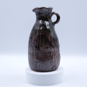 Puede incluir: Una jarra de cerámica marrón oscuro hecha a mano, con un asa pequeña y un pico. La jarra tiene un diseño acanalado vertical texturizado y se asienta sobre un pedestal blanco. El esmalte tiene un acabado brillante.
