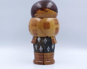 May include: A wooden kokeshi doll featuring a dark brown rounded head, a light brown torso with a tan plaid pattern, and a dark brown lower body decorated with small white floral motifs, finished with light brown carved wooden boots.