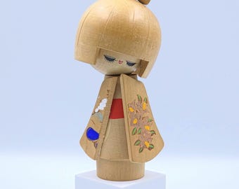 Op de afbeelding: Een houten Kokeshi-pop met een ronde kop en een rode strik. De pop draagt een kledingstuk in kimono-stijl met bloemen- en wolkenmotieven. De pop staat op een witte kubus.