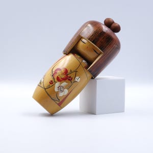 May include: A wooden Kokeshi doll with a brown head and a yellow body. The doll's body is decorated with red and white floral designs. The doll is resting on a white cube against a white background.