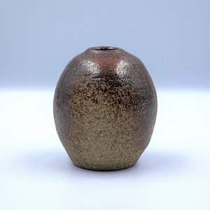 May include: A small, handcrafted ceramic vase with a rounded, oval shape. The vase has a dark brown, textured surface with a gradient of colors, transitioning from a lighter tan at the base to a darker, reddish-brown at the top. The opening is a small, circular hole.