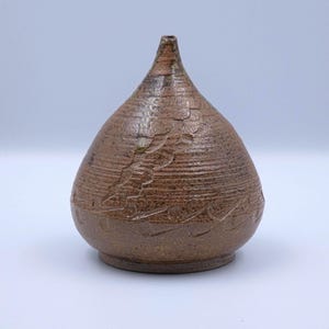 May include: A handcrafted brown ceramic vase with a textured surface and a unique, teardrop shape. The vase features carved floral patterns and horizontal lines. The vase is approximately 15 cm tall.