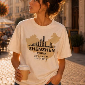 May include: A cream-colored short-sleeve t-shirt featuring a tan map silhouette of China. Centered on the map is a black skyline illustration of Shenzhen, with the text SHENZHEN CHINA and the coordinates 22° 32' 42" N 114° 3' 54" E printed below.