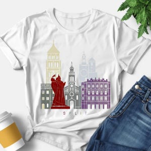 Peut inclure: T-shirt blanc avec un motif coloré de l'horizon de Split. Le design comprend une statue rouge, des bâtiments en jaune, gris et violet, et le mot "SPLIT" en blanc.