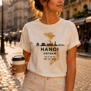 May include: A cream-coloured short-sleeve t-shirt featuring a brown watercolour map of Vietnam, a black skyline silhouette, and the text HANOI VIETNAM 21° 2' 0" N 105° 51' 0" E printed in black ink on the centre of the chest area.