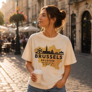Peut inclure: Un t-shirt à manches courtes de couleur crème avec un graphique noir représentant la ligne d'horizon de Bruxelles sur une forme de carte dorée style aquarelle. Le texte indique BRUSSELS BELGIUM avec les coordonnées 50 50 47 N 4 21 8 E.