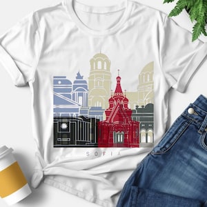 May include: A white t-shirt featuring a graphic print of the Sofia city skyline. The design includes stylized architectural silhouettes in shades of red, blue, beige, and black, with the word SOFIA printed in small black letters at the bottom.