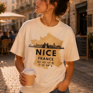 Puede incluir: Una camiseta de manga corta color crema con un mapa de Francia en tono bronceado y una silueta negra del horizonte de Niza. El texto NICE FRANCE aparece en letras negras, seguido de las coordenadas 43° 42' 12" N y 7° 15' 59" E en negrita.