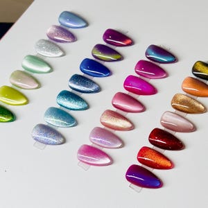 May include: A collection of artificial nails in various colors, arranged in rows. The nails display a range of hues, including green, yellow, pink, blue, and purple, with glitter and iridescent effects. Each nail is mounted on a clear plastic tab.