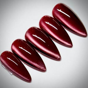 May include: Five almond-shaped artificial nails with a deep burgundy color and a glossy finish. The nails are arranged in a row, showcasing their elongated shape and reflective surface. The color shifts slightly with the light.