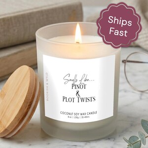 May include: A frosted glass candle with a lit wick and a light wood lid. The white label features the text Smells Like... PINOT & PLOT TWISTS, BOOKISH & WYLD, and COCONUT SOY WAX CANDLE 8 oz | 230g | 35-45hrs. The candle emits a warm, bright flame.