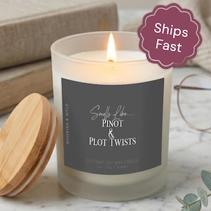 May include: A frosted glass candle with a lit wick and a bamboo lid. The dark gray label reads: Smells Like... PINOT & PLOT TWISTS, BOOKISH & WYLD, COCONUT SOY WAX CANDLE, 8 oz | 230g | 35-45hrs. The candle is positioned next to its wooden lid.