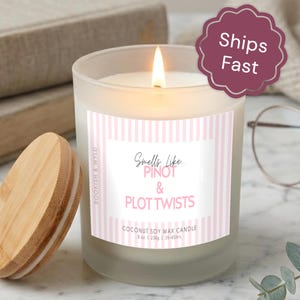 May include: A frosted glass candle with a lit wick and a light wood lid. The label features pink vertical stripes and text that reads: Smells Like... PINOT & PLOT TWISTS, BOOKISH & WYLD, COCONUT SOY WAX CANDLE, 8 oz | 230g | 35-45hrs.