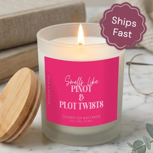 May include: A frosted glass candle with a bright pink label featuring the white text Smells Like PINOT & PLOT TWISTS, Coconut Soy Wax Candle, 8 oz, 230g, 35-45hrs. The candle has a lit wick and is accompanied by a round, light-colored wooden lid.