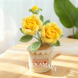 May include: A crocheted potted plant featuring two large yellow roses and one small yellow bud with green leaves. The pot is beige with a scalloped top edge and a rectangular tan label that reads Hand Made with Love and the text FLORA AMARA.