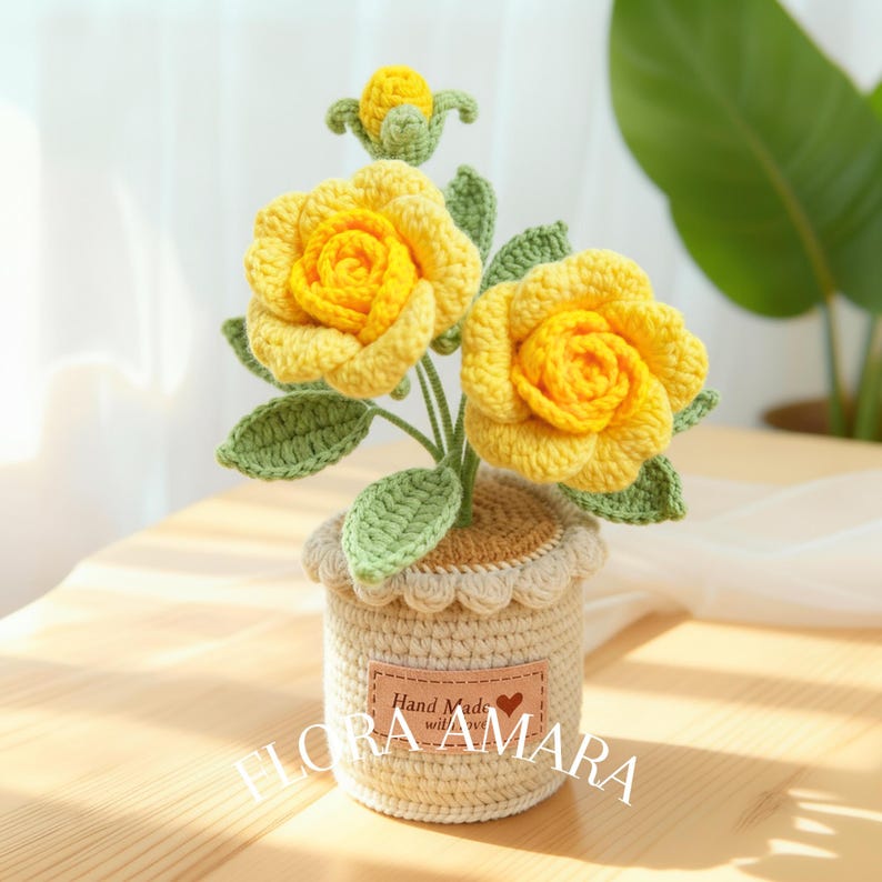 May include: A crocheted potted plant featuring two large yellow roses and one small yellow bud with green leaves. The pot is beige with a scalloped top edge and a rectangular tan label that reads Hand Made with Love and the text FLORA AMARA.