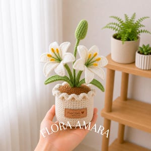 Puede incluir: Una planta de lirio tejida a ganchillo en una maceta color crema con un borde superior ondulado. La planta presenta dos flores de lirio blancas con estambres amarillos, tallos verdes y un capullo verde. Una etiqueta marrón dice Hand Made with Love.