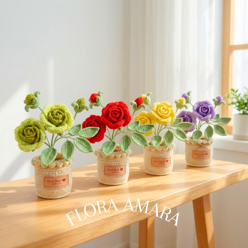 May include: Four crocheted rose arrangements in small, beige, textured pots. Each pot features a label reading "Hand Made" with a red heart. The roses are in shades of light green, red, yellow, and purple, each with green leaves and buds.