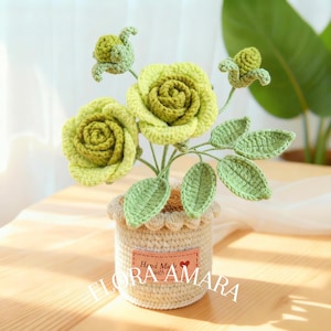 May include: A crocheted potted plant featuring two blooming roses and two buds in shades of light and dark green. The flowers have textured petals and green leaves on stems. The pot is beige with a scalloped rim and a label reading Hand Made with love.