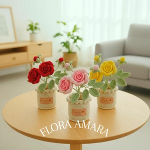 May include: Three crocheted rose arrangements in cream-coloured pots. Each pot features a small brown tag with the text Hand Made with love. The roses are red, pink, and yellow, accompanied by green crocheted leaves and small buds on stems.