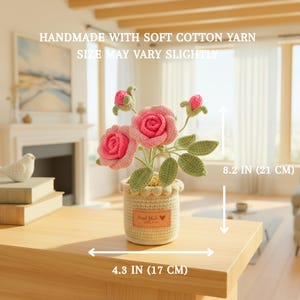 May include: A crochet rose plant in a cream-coloured pot with a label reading Hand Made. The plant features pink roses, green leaves, and two buds. It measures 21 cm in height and 17 cm in width. Text states: HANDMADE WITH SOFT COTTON YARN.