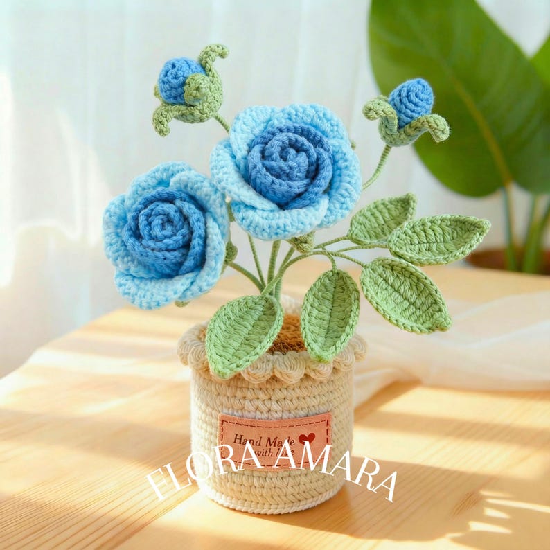 May include: A crocheted flower arrangement featuring two large blue roses and two small blue buds with light green leaves. The flowers are set in a cylindrical beige crocheted pot with a scalloped top edge and a label reading Hand Made with love FLORA AMARA.